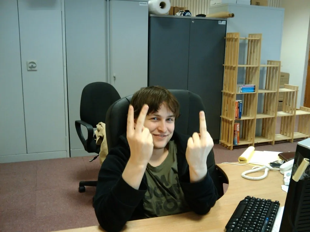 A young man at a desk in a server room, raising both middle fingers